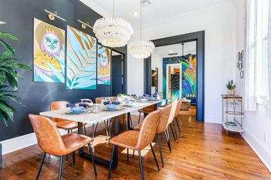 Bright modern dining room with a long marble table set for eight, tan leather chairs, two white tiered chandeliers, dark accent wall with three-panel colorful tropical artwork, hardwood floors, indoor plants, and an open doorway revealing a vibrant mural in the next room.