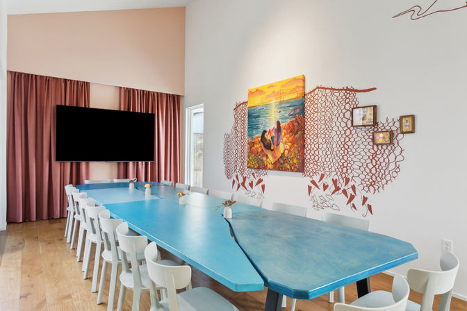 Bright coastal-themed meeting room with long turquoise conference table surrounded by white chairs, sunset seaside painting framed by red net mural on a white wall, large black flat-screen TV and blush curtains