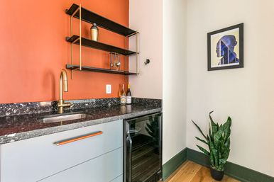 Modern kitchenette with orange accent wall, black marble countertop and brass faucet, black-and-gold open shelving, built-in wine fridge, framed portrait on white wall, and potted snake plant on hardwood floor.