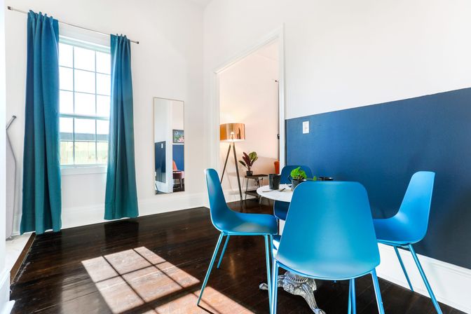 Sun-dappled modern dining nook with four bright blue chairs around a white round table on dark hardwood floors, teal curtains framing a large window, navy half-wall accent, full-length mirror and a doorway revealing a tripod lamp and potted plant.