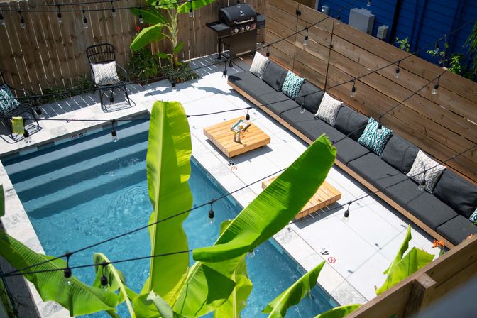 Aerial view of a compact modern backyard pool oasis with turquoise water, concrete deck, long gray cushioned bench with patterned pillows, wooden coffee tables, banana-leaf plants, hanging string lights and a barbecue grill.