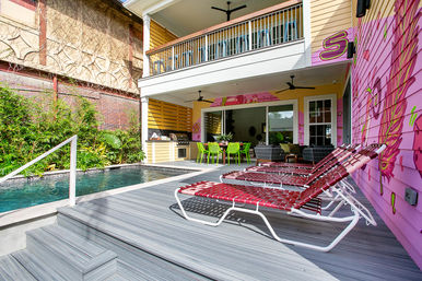 Vibrant tropical backyard with private plunge pool, gray composite deck and four red woven lounge chairs beside a pink muraled exterior wall, covered patio with outdoor kitchen and green dining chairs under a second-story balcony.