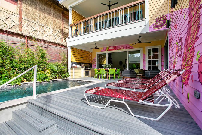 Vibrant tropical backyard with private plunge pool, gray composite deck and four red woven lounge chairs beside a pink muraled exterior wall, covered patio with outdoor kitchen and green dining chairs under a second-story balcony.
