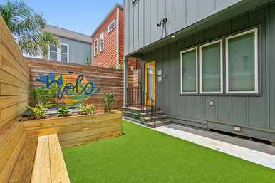 Small urban townhouse courtyard with bright yellow door, gray siding, artificial turf, raised wooden planter and colorful "hello" mural on a wooden privacy fence
