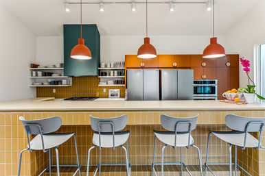 Modern kitchen with breakfast bar: mustard-tiled island, four pale-blue bar stools, three orange pendant lights, stainless steel fridge and built-in ovens, open shelving and orchid
