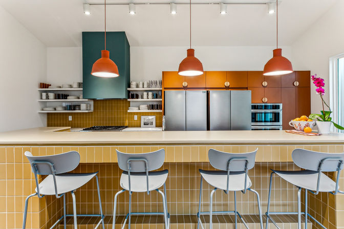 Modern kitchen with breakfast bar: mustard-tiled island, four pale-blue bar stools, three orange pendant lights, stainless steel fridge and built-in ovens, open shelving and orchid