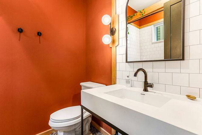 Cozy modern powder room interior with burnt orange accent wall, white rectangular vanity sink and subway tile backsplash, matte black faucet and fixtures, arched mirror and globe wall sconces