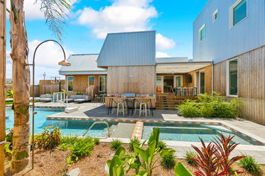 Sunny modern backyard oasis with rectangular pool and spa, wooden-clad contemporary house, outdoor bar and lounge seating, palm trees and tropical landscaping under a bright blue sky.