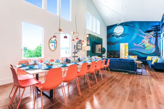 Bright open-plan dining and living room with a long white table set for many, row of coral chairs, blue velvet sofas, large colorful ocean-themed mural, vaulted windows and warm hardwood floors.