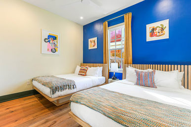 Sunny vacation rental bedroom with two queen beds, bold cobalt accent wall, wood-slat headboards, mustard curtains framing a window, colorful woven throws and patterned pillows on hardwood floors.