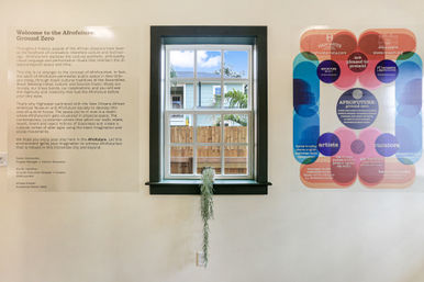 Bright art gallery wall with a centered mullioned window looking out to a wooden fence and light-blue house, a trailing potted plant hangs from the sill, large exhibit text panel to the left and a vibrant circular infographic poster to the right.