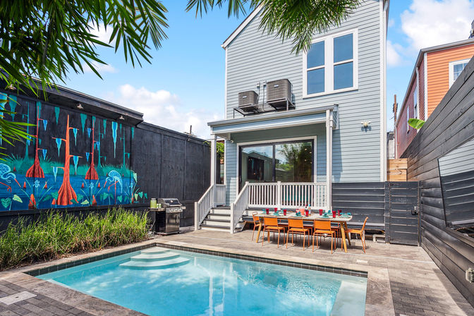 Backyard pool and patio at a modern two-story townhouse — rectangular plunge pool, poolside dining table with orange chairs, colorful mural on black fence, gas grill and tropical plants.