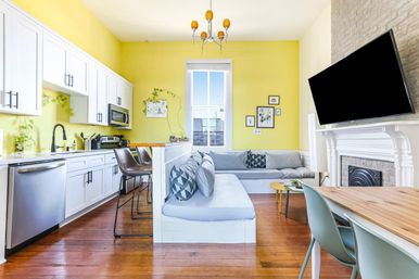Sunny open-plan apartment kitchen and living room with bright yellow walls, white cabinets, stainless appliances, built-in banquette seating, hardwood floors, and wall-mounted TV