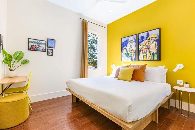 Bright modern bedroom with bold yellow accent wall, wooden platform bed with white linens and colorful throw pillows, vibrant framed art, hardwood floors, small desk and potted plant — ideal vacation rental room.