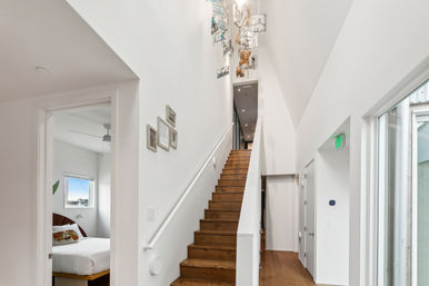 Sunlit modern home interior with white walls and tall vaulted ceiling, wooden staircase with white handrail, contemporary hanging glass chandelier, bedroom doorway visible on left and glass exterior door on right.