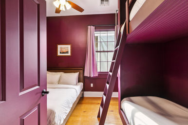 Cozy plum-painted bedroom with a double bed, laddered bunk bed, hardwood floors, ceiling fan and a curtained window.
