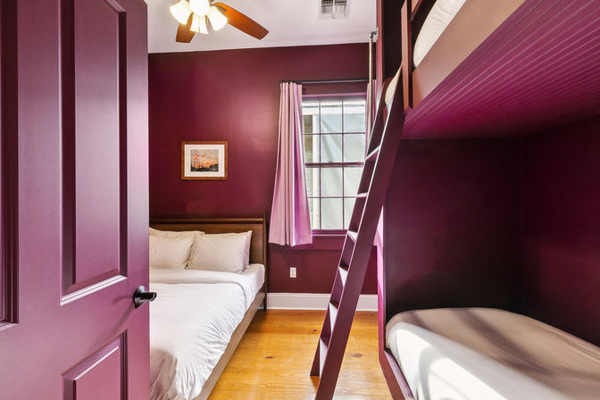 Cozy plum-painted bedroom with a double bed, laddered bunk bed, hardwood floors, ceiling fan and a curtained window.