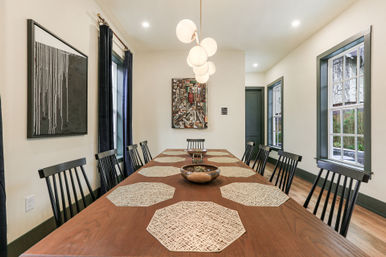 Modern residential dining room with long wooden table topped with woven octagonal placemats, black spindle-back chairs, globe pendant lights, abstract wall art and tall windows with dark trim.