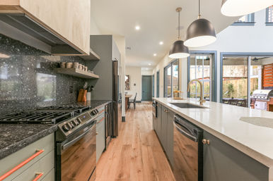 Bright modern open-plan kitchen with a long white island, pendant lights, stainless appliances, black stone countertops, wood floors, and sliding glass doors to a patio grill.