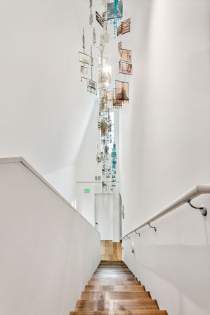 Bright modern stairwell with wooden steps and white walls, featuring a tall hanging glass mobile art chandelier with colorful panels