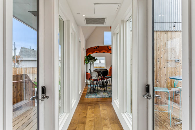 Modern narrow hallway with hardwood floors and floor-to-ceiling glass doors leading to a cozy dining nook with high stools, a leafy plant and orange retro accent wall; outdoor wooden deck and turquoise bistro chairs visible through the glass.