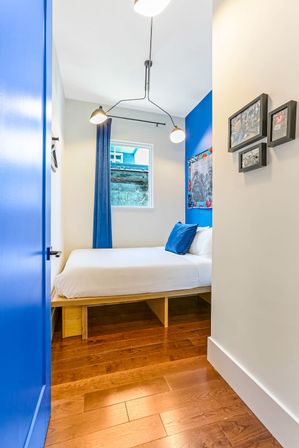 Cozy modern small bedroom with cobalt blue door and accent wall, platform single bed with white linens and blue pillow, hardwood floors, window with blue curtain and contemporary pendant light.