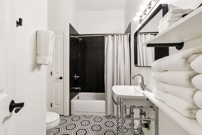 Modern black-and-white bathroom with hexagon mosaic floor, white tub and herringbone black-tile shower, white sink with exposed chrome pipes, black-framed mirror, shower curtain and stacked fluffy towels on open shelves.