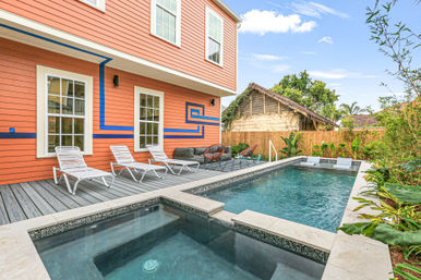 Backyard pool and spa beside an orange two-story house with blue geometric trim, gray deck loungers, tropical plants and a wooden fence.