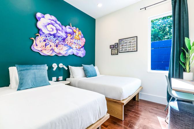 Bright boutique-style twin guest room with teal accent wall and whimsical purple cloud mural above two white beds with blue pillows, hardwood floors, window with green view, desk and potted plant.