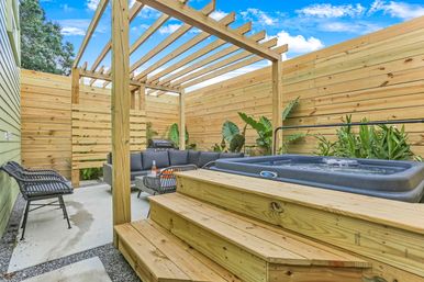 Cozy sunny backyard patio with wooden pergola and slatted privacy fence, raised hot tub with wooden steps, gray outdoor sectional and wicker chairs around a coffee table, barbecue grill and tropical plants.