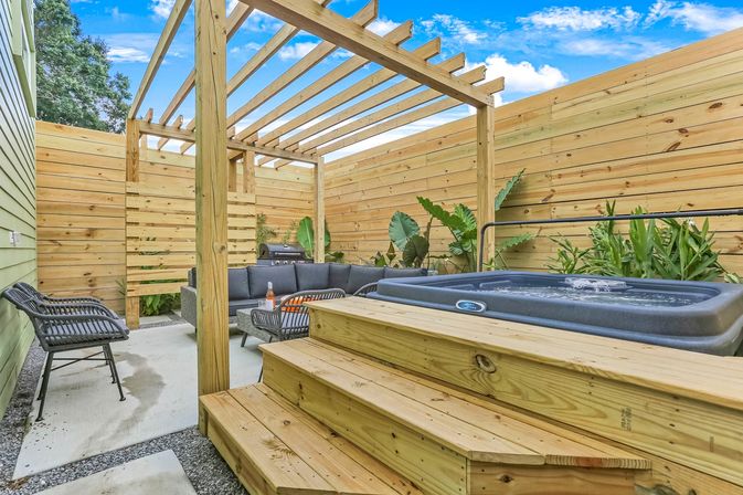 Cozy sunny backyard patio with wooden pergola and slatted privacy fence, raised hot tub with wooden steps, gray outdoor sectional and wicker chairs around a coffee table, barbecue grill and tropical plants.