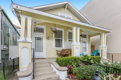 Pale yellow Craftsman bungalow with wide covered front porch, hanging planters and wooden swing, teal chairs, beige steps and manicured front garden.
