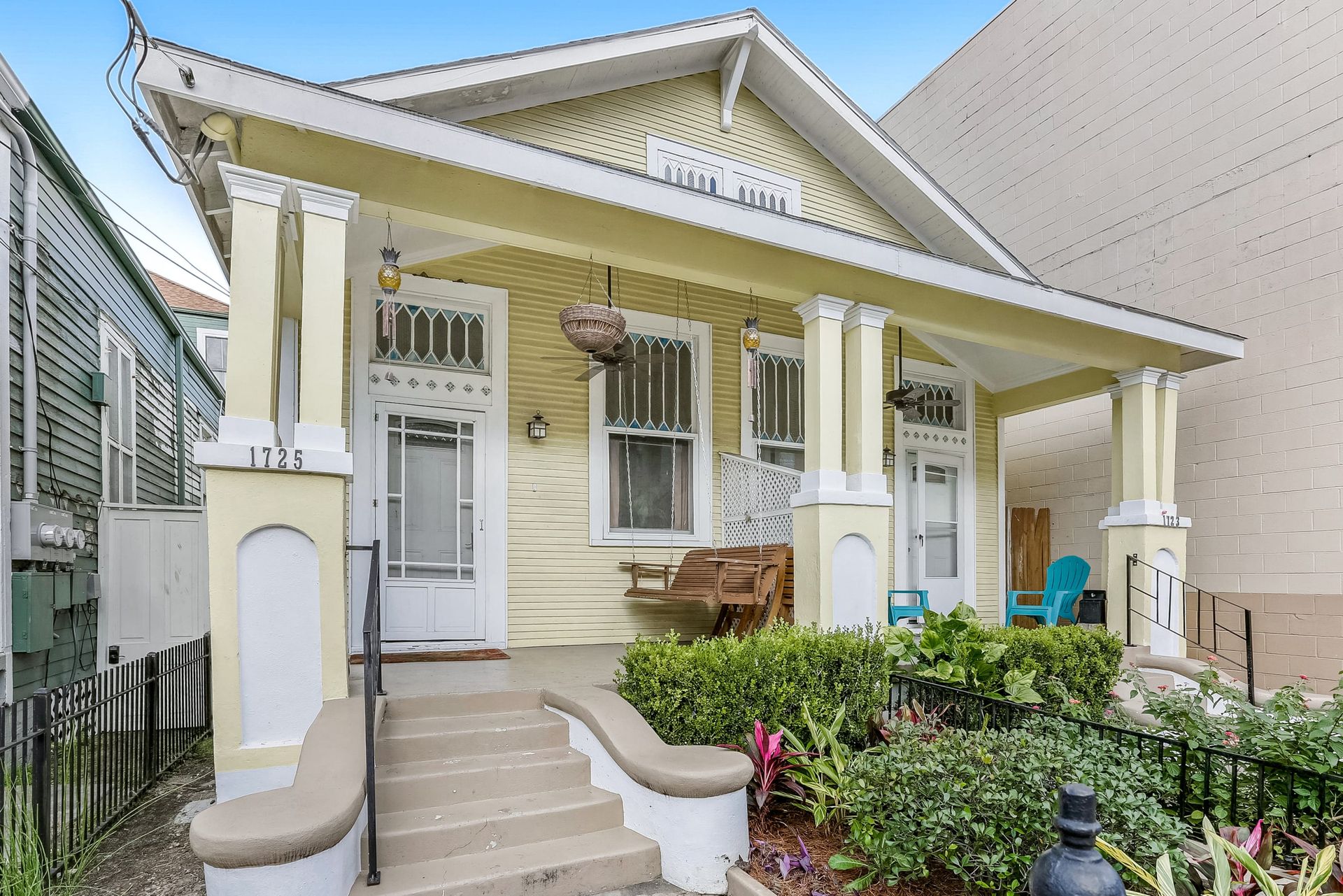Pale yellow Craftsman bungalow with wide covered front porch, hanging planters and wooden swing, teal chairs, beige steps and manicured front garden.