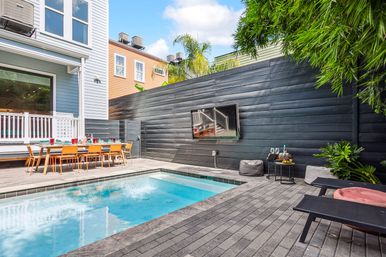 Sunny urban townhouse backyard with a rectangular plunge pool, long outdoor dining table with orange chairs, black wood privacy wall with wall-mounted TV, lounge chairs and tropical plants.