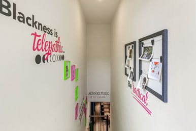 Narrow modern creative stairwell with bold wall lettering “Blackness is Telepathic,” neon pink and green picture frames along the left wall and clipped photo displays with a pink “Be radical” script on the right, looking down to a lower level.