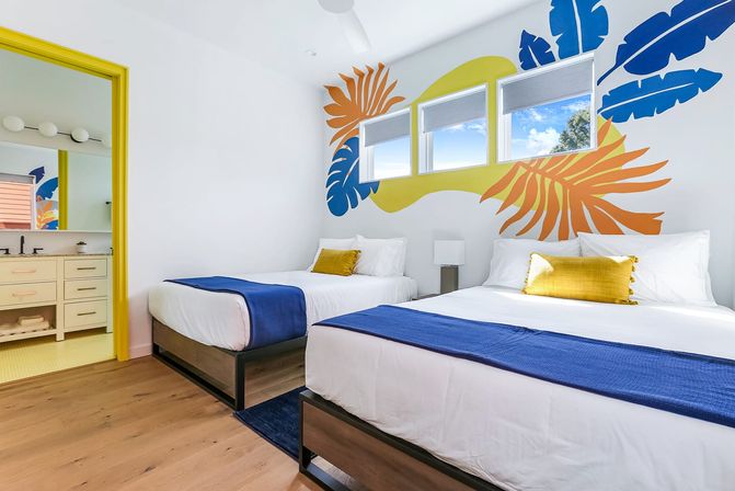 Bright coastal-themed guest room with two beds, colorful tropical leaf mural, blue throws, yellow accent pillow and trim, hardwood floor and sunny windows