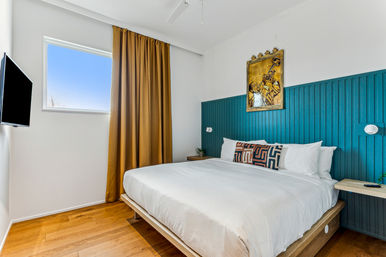 Bright modern bedroom with a king bed and white linens, teal vertical-paneled headboard, patterned accent pillow, mustard curtains, wood floors, wall-mounted TV and trumpet artwork above the bed.
