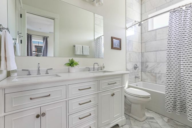 Clean, bright white bathroom with a double-sink vanity, marble-tiled tub/shower, striped shower curtain, chrome fixtures, large mirror, and small potted plant.