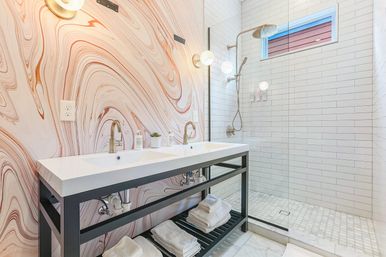 Bright modern bathroom with double-sink vanity, marble-swirl accent wall, glass walk-in subway-tiled shower with rainfall head and folded towels on open shelf.