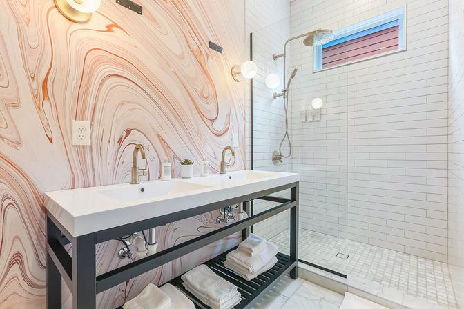 Bright modern bathroom with double-sink vanity, marble-swirl accent wall, glass walk-in subway-tiled shower with rainfall head and folded towels on open shelf.