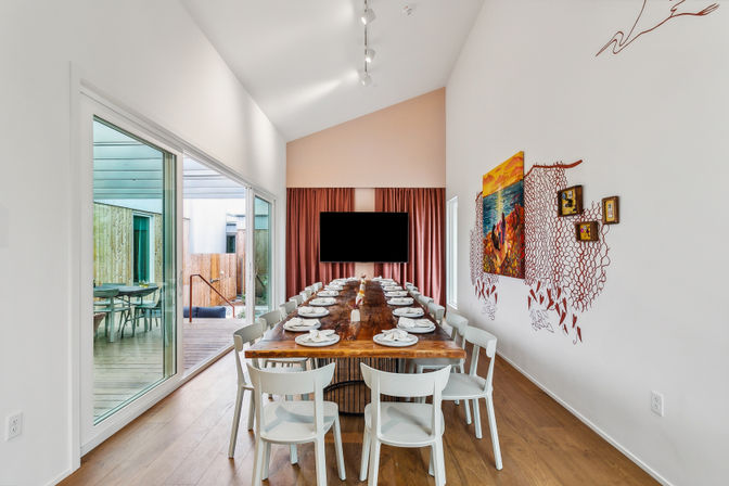 Modern dining room with a long reclaimed-wood table set for a large group, white chairs on hardwood floors, sliding glass doors to an outdoor deck, pink curtains, colorful wall art and a wall-mounted TV.