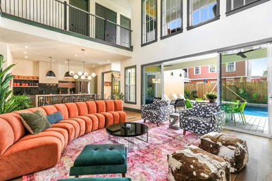 Bright open-concept living room with double-height ceiling, curved terracotta velvet sofa, patterned armchairs, colorful area rug, kitchen island with pendant lights, and sliding glass doors opening to a backyard pool and patio.