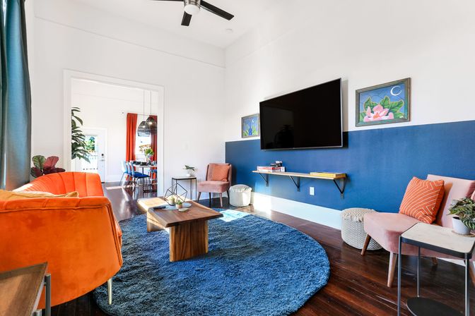Sunlit modern living room with navy half-wall and wall-mounted TV, orange velvet sofa, round blue shag rug, wooden coffee table and pink accent chairs, open-plan dining area visible through doorway.