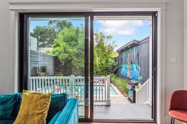 Sliding glass door view of a modern backyard: teal sofa with yellow pillow inside, small deck and railing leading to a narrow pool, outdoor grill, vibrant mural on the fence, and lush bamboo.
