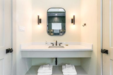 Spa-like modern bathroom with white floating vanity and undermount sink, matte-black faucet, rounded rectangular mirror flanked by warm wall sconces, soap dispenser and neatly folded white towels on the lower shelf.