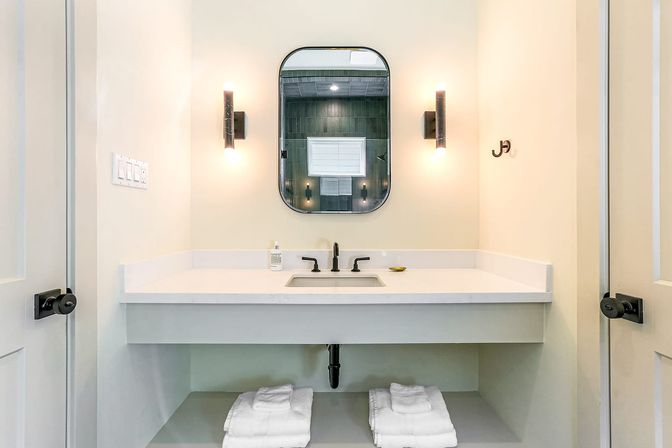 Spa-like modern bathroom with white floating vanity and undermount sink, matte-black faucet, rounded rectangular mirror flanked by warm wall sconces, soap dispenser and neatly folded white towels on the lower shelf.