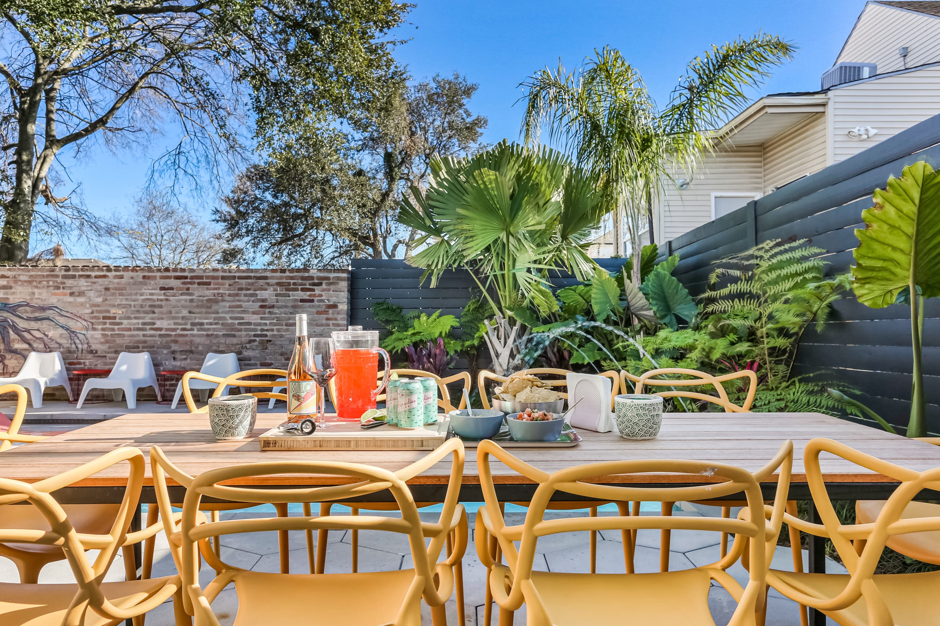 Sunny backyard patio with a long wooden outdoor dining table, yellow modern chairs, pitcher of red punch, wine bottle, snacks and canned drinks, surrounded by tropical plants, brick wall and blue sky.