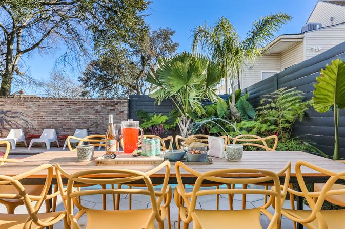 Sunny backyard patio with a long wooden outdoor dining table, yellow modern chairs, pitcher of red punch, wine bottle, snacks and canned drinks, surrounded by tropical plants, brick wall and blue sky.