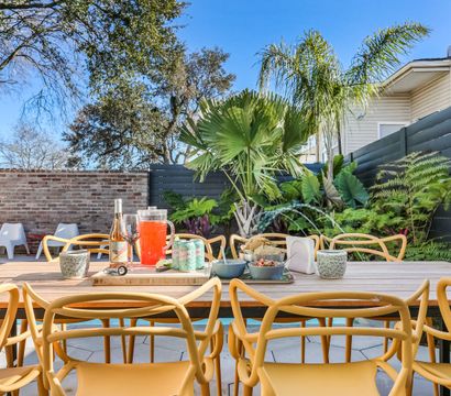 Sunny backyard patio with a long wooden outdoor dining table, yellow modern chairs, pitcher of red punch, wine bottle, snacks and canned drinks, surrounded by tropical plants, brick wall and blue sky.