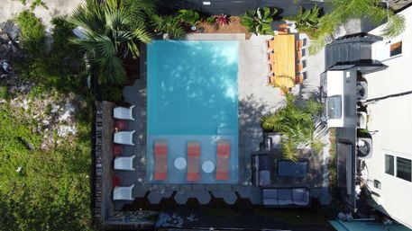Aerial drone view of a turquoise rectangular backyard pool with three submerged lounge chairs, a row of sunbeds, palm trees, patio seating and a wooden outdoor dining table.